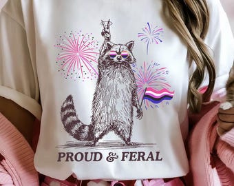 May include: White t-shirt with a raccoon graphic wearing sunglasses, holding a flag, and fireworks. The text "PROUD & FERAL" is below the raccoon. The person is wearing pink heart-patterned shorts and a pink sweater.