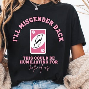 May include: Black t-shirt featuring the text "I'll Misgender Back" in pink, and a pink and white reverse card graphic. The phrase "This could be humiliating for both of us" is printed below the card.