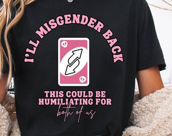 Puede incluir: Camiseta negra con el texto rosa "I'll Misgender Back" y un gráfico de carta de reversa rosa y blanca. La frase "This could be humiliating for both of us" está impresa debajo.