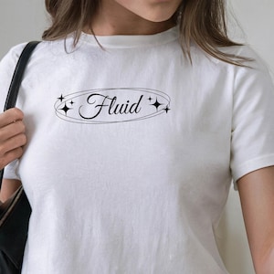 Puede incluir: Top corto blanco con la palabra "Fluid" en fuente negra, enmarcada por un óvalo negro y estrellas. La camiseta se combina con vaqueros de mezclilla azul claro. Un bolso negro cuelga del hombro.
