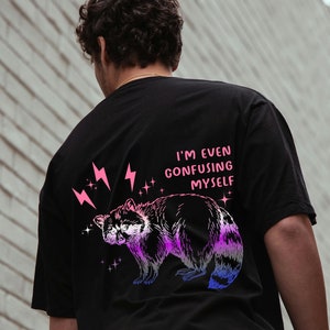 Puede incluir: Camiseta negra con un gráfico de un mapache con acentos rosa y azul. El texto "I'm even confusing myself" está impreso en la camiseta.