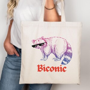 May include: Cream tote bag with a purple and pink raccoon wearing pixelated sunglasses. The word "Biconic" is printed in red script below the raccoon. The bag has two handles and is held by a person.