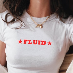 Puede incluir: Camiseta blanca con la palabra "FLUID" en rojo, con una estrella roja a cada lado. La camiseta es de manga corta y entallada. Se ve un collar dorado.