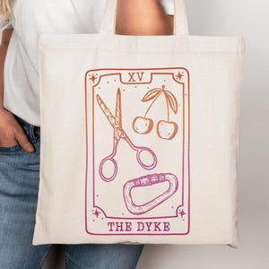May include: Cream-colored tote bag featuring a tarot card design with scissors, cherries, and a carabiner. The card reads "XV" at the top and "THE DYKE" at the bottom, with an orange to purple gradient. The bag is held by a person.