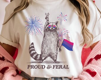 May include: White t-shirt featuring a raccoon wearing sunglasses and holding a flag with the bisexual pride colors. The raccoon is standing in front of fireworks. The text "PROUD & FERAL" is printed below the raccoon.