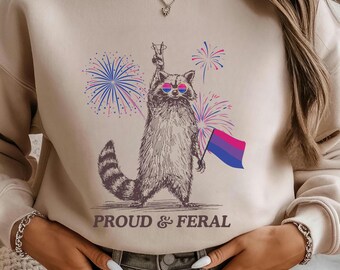May include: Beige sweatshirt featuring a raccoon wearing sunglasses and holding a bisexual pride flag. The raccoon is surrounded by fireworks in blue, pink, and purple. The text "PROUD & FERAL" is printed below the raccoon.