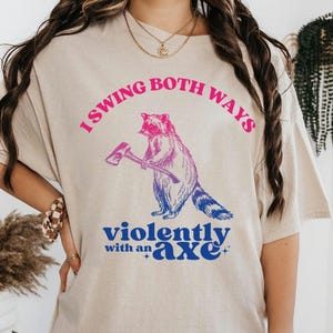 Puede incluir: Camiseta beige con el texto "I swing both ways violently with an axe". El gráfico presenta un mapache sosteniendo un hacha, con un degradado de color rosa y azul.
