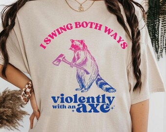 May include: Beige t-shirt with the text "I swing both ways violently with an axe." The graphic features a raccoon holding an axe, with pink and blue gradient coloring.
