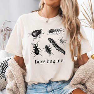 Può includere: T-shirt color crema con illustrazioni nere di vari insetti, tra cui uno scarabeo, una formica e un ragno. La frase "boys bug me" è stampata sotto. La maglietta ha una vestibilità ampia ed è in cotone.