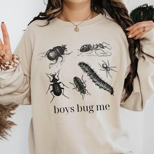 May include: Beige sweatshirt featuring black illustrations of various insects, including a beetle, ant, and spider. The phrase "boys bug me" is printed in a black, sans-serif font below the insects. The sweatshirt has a crew neck and long sleeves.