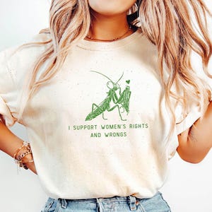Può includere: Felpa color crema con una grafica verde di due mantidi religiose che tengono un cuore. Il testo "I SUPPORT WOMEN'S RIGHTS AND WRONGS" è stampato in verde sotto.