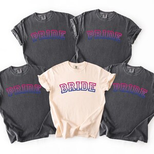 May include: Five t-shirts with the word "PRIDE" or "BRIDE" in a bi-pride color scheme. Four shirts are dark gray, and one is cream-colored. The text is in a bold, arched font, with the colors pink, blue, and white.