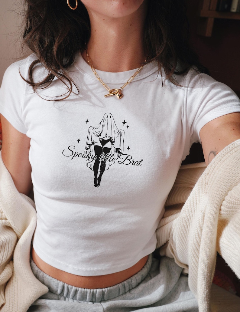May include: White crop top with a black graphic of a ghost figure and the text "Spooky little Brat". The design includes stars and a stylized illustration. The shirt is a casual, everyday wear item.