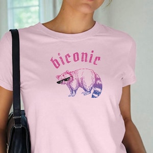 Puede incluir: Camiseta rosa claro con un mapache con gafas de sol y la palabra "Biconic" encima en un degradado rosa y morado. La ilustración del mapache también está en un degradado rosa y morado.