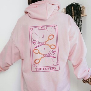 Könnte beinhalten: Hellrosa Kapuzenpullover mit einem Tarotkarten-Design auf dem Rücken. Die Karte zeigt zwei Scherenpaare mit herzförmigen Klingen, die Zahl VI und die Worte "THE LOVERS" in Rosa und Lila.