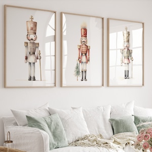 Puede incluir: Tres acuarelas de soldados cascanueces en diferentes poses y colores. Las pinturas están enmarcadas en madera natural y colgadas en una pared blanca sobre un sofá blanco.