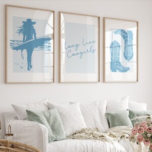 May include: Three framed prints with blue illustrations on a white wall above a white couch with light blue pillows. The prints feature a woman surfing, the text "long live cowgirls", and a pair of cowboy boots. A bouquet of pink flowers sits on a wooden coffee table in front of the couch.
