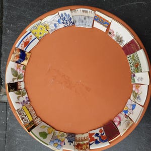 May include: A circular terracotta clay saucer featuring a decorative border made of various broken ceramic pottery shards. The shards display diverse patterns including floral motifs, geometric shapes, and blue and white designs arranged around the rim.