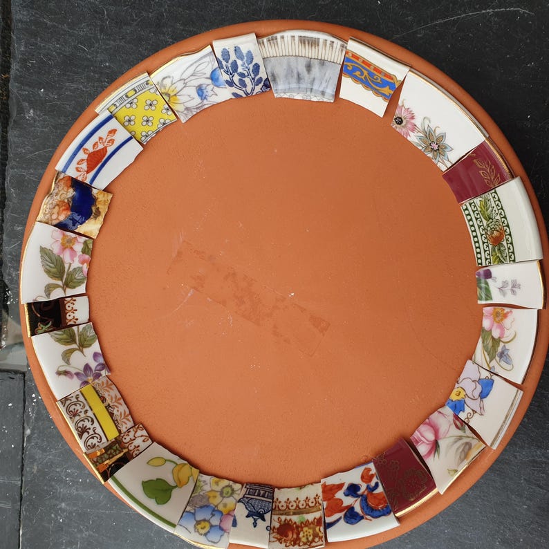 May include: A circular terracotta clay saucer featuring a decorative border made of various broken ceramic pottery shards. The shards display diverse patterns including floral motifs, geometric shapes, and blue and white designs arranged around the rim.