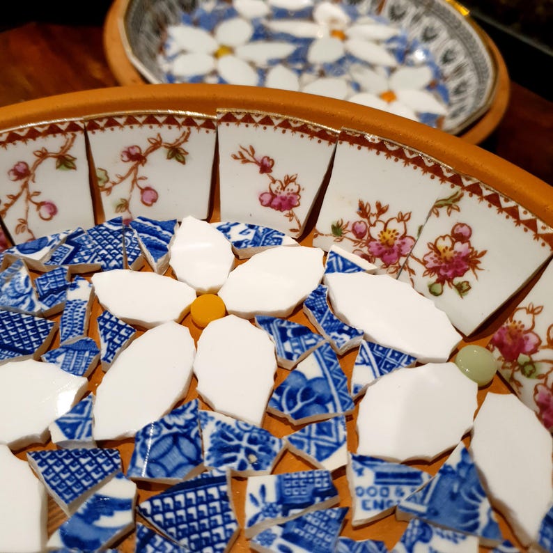 May include: A circular terracotta bowl featuring a mosaic design made of broken ceramic pieces. The rim is lined with white porcelain shards decorated with pink floral patterns, while the center displays white petal shapes and blue patterned shards.