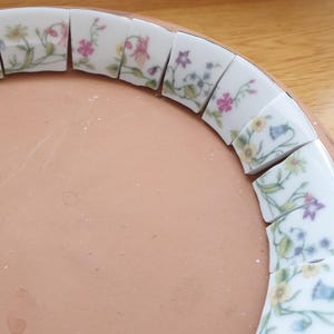 May include: A circular terracotta base features a border of curved white ceramic tiles arranged in a ring. Each tile displays a delicate floral pattern with green stems, leaves, and small blossoms in shades of pink, yellow, purple, and blue.