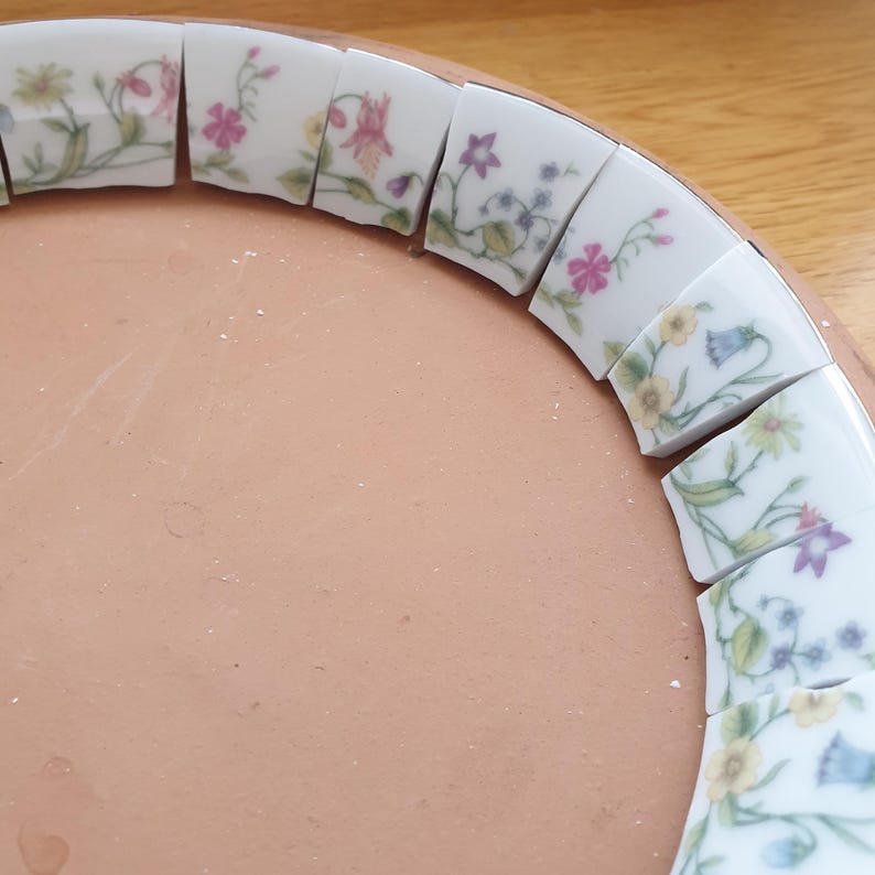 May include: A circular terracotta base features a border of curved white ceramic tiles arranged in a ring. Each tile displays a delicate floral pattern with green stems, leaves, and small blossoms in shades of pink, yellow, purple, and blue.