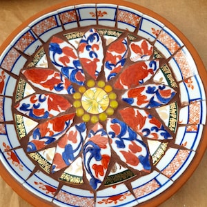 May include: A circular mosaic plate featuring a central yellow floral tile surrounded by small yellow glass beads. The design includes radiating petal-shaped ceramic shards in white, blue, and orange, set against dark brown grout within a terracotta rim.