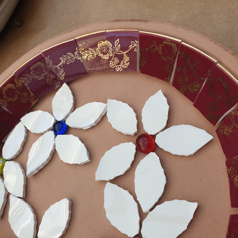 May include: A circular terracotta base featuring a mosaic design. The border consists of deep red ceramic tiles with gold floral patterns. The center displays white petal-shaped tiles arranged into flower motifs with blue and red glass centers.
