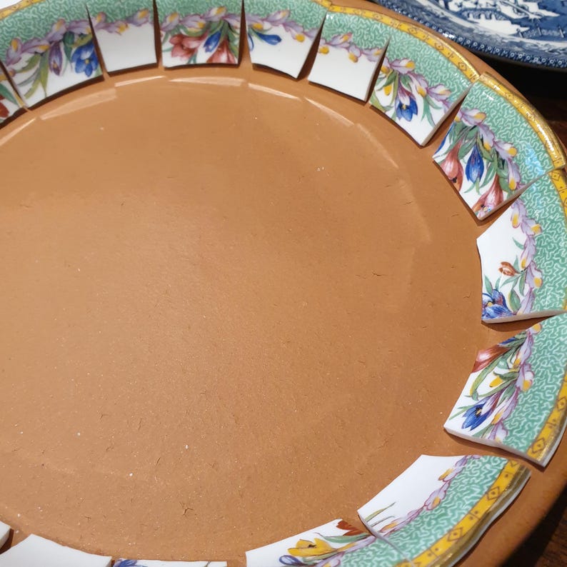 May include: A circular terracotta plate featuring a decorative border made of individual ceramic shards. Each shard displays a green patterned background with colorful floral motifs in shades of blue, yellow, and red, finished with a gold rim.
