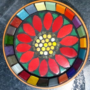 May include: A red mosaic flower with a yellow center surrounded by a ring of colorful mosaic tiles.