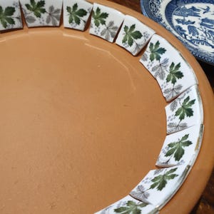 May include: A terracotta clay saucer featuring a decorative border of white ceramic tile fragments. Each tile is adorned with a repeating pattern of green and grey maple leaves and small clusters of berries arranged along the inner rim.