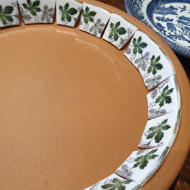 May include: A terracotta clay saucer featuring a decorative border of white ceramic tile fragments. Each tile is adorned with a repeating pattern of green and grey maple leaves and small clusters of berries arranged along the inner rim.