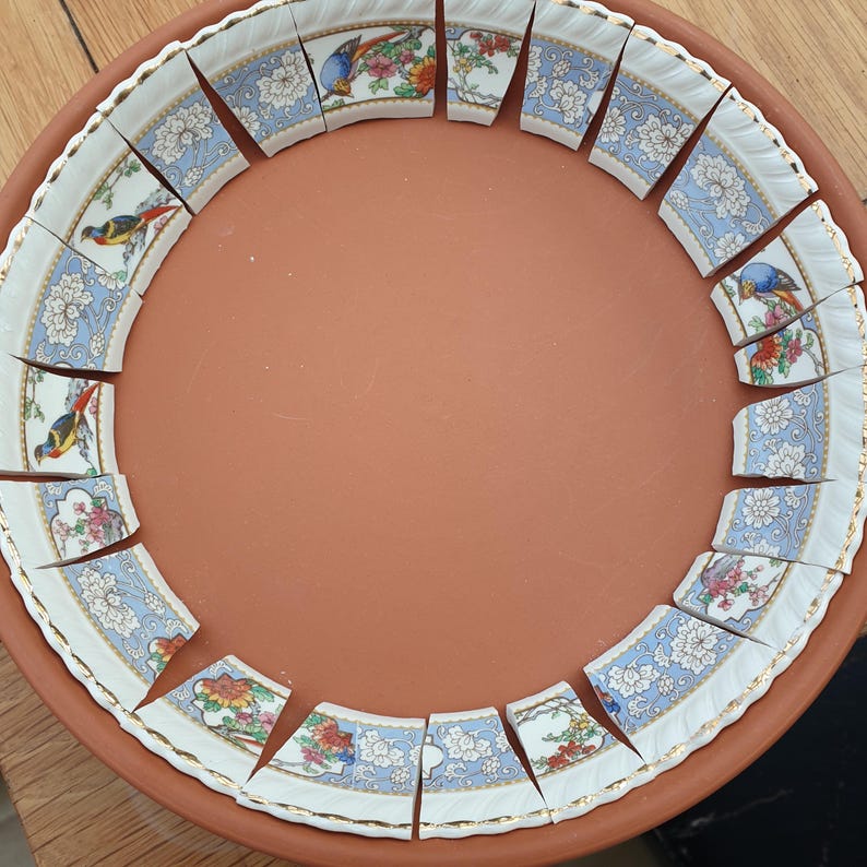 May include: A circular terracotta tray containing a series of curved ceramic shards arranged along the inner rim. The shards feature a white background with blue floral patterns, gold-trimmed edges, and colorful depictions of small birds.