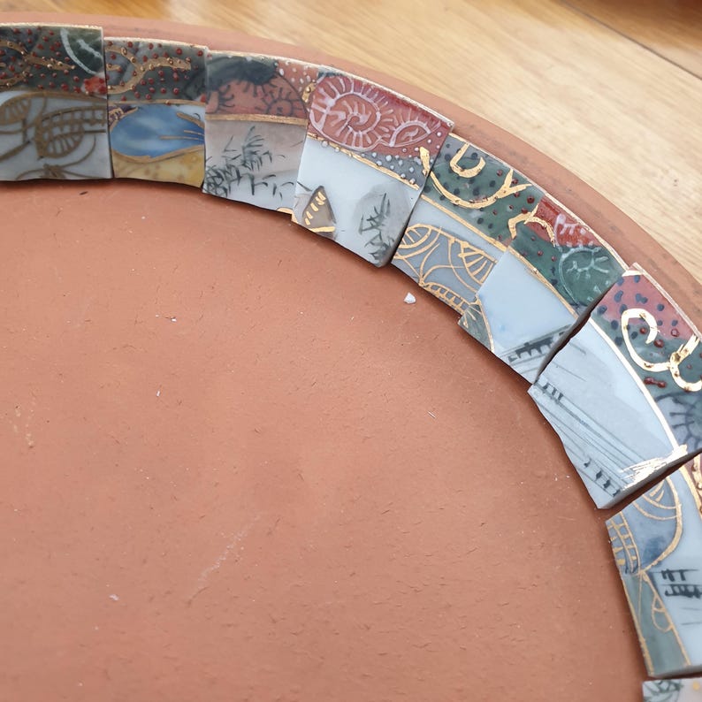May include: A curved arrangement of ceramic mosaic tiles featuring various patterns in blue, red, green, and white with gold-colored accents. The tiles are placed along the inner edge of a smooth, circular, terracotta-colored clay base.