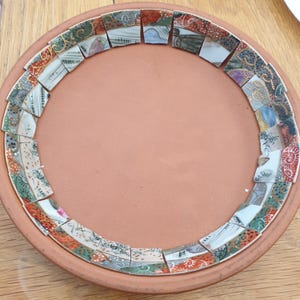 May include: A round terracotta plate featuring a decorative border made of small, colorful ceramic shards. The shards display various patterns in shades of orange, green, blue, and white, arranged in a circular mosaic pattern around the center.