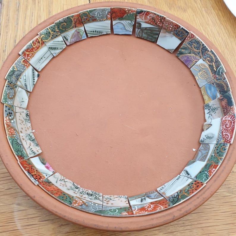 May include: A round terracotta plate featuring a decorative border made of small, colorful ceramic shards. The shards display various patterns in shades of orange, green, blue, and white, arranged in a circular mosaic pattern around the center.