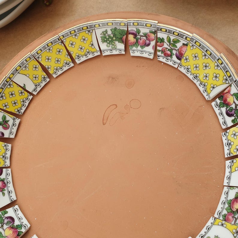 May include: A circular terracotta base features a decorative border made of curved ceramic shards. The shards alternate between a yellow pattern with white flowers and white segments displaying colorful illustrations of grapes, apples, and plums.