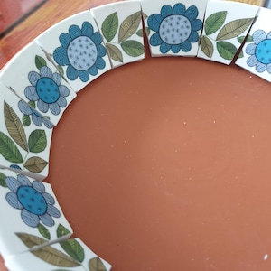 May include: A terracotta bowl featuring a decorative inner rim composed of white ceramic segments. Each segment displays a blue flower with a dotted center and green and brown leaves with fine black line details arranged in a repeating pattern.