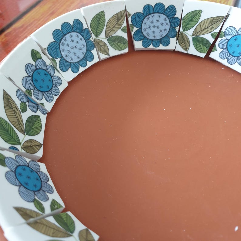 May include: A terracotta bowl featuring a decorative inner rim composed of white ceramic segments. Each segment displays a blue flower with a dotted center and green and brown leaves with fine black line details arranged in a repeating pattern.