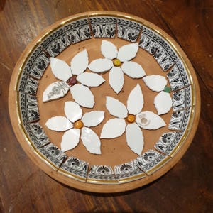 May include: A round terracotta plate featuring a mosaic design. The border consists of black and white patterned ceramic shards with gold trim. The center displays three white flower shapes made from ceramic pieces with small colored centers.