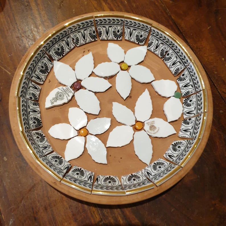 May include: A round terracotta plate featuring a mosaic design. The border consists of black and white patterned ceramic shards with gold trim. The center displays three white flower shapes made from ceramic pieces with small colored centers.