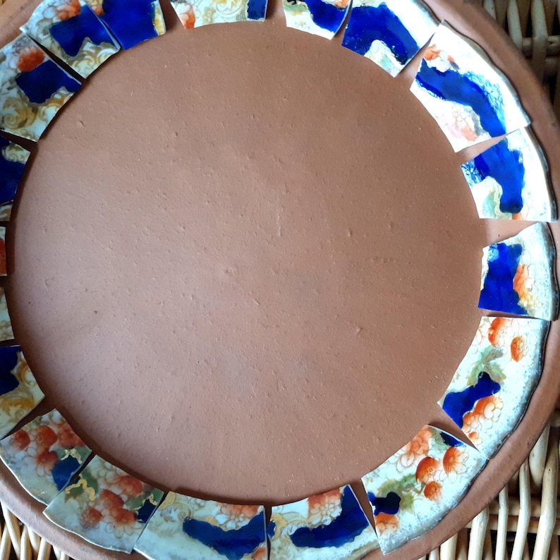 May include: A circular terracotta plate featuring a decorative border made of broken ceramic shards. The shards are white with patterns of deep blue and orange floral designs, arranged in a ring around the smooth, solid brown clay center.