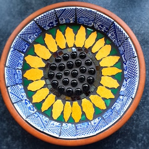 May include: A mosaic bowl with a sunflower design. The centre of the sunflower is made of black glass beads, surrounded by yellow petals. The rim is decorated with blue and white patterned tiles, and the outer edge is a warm brown.