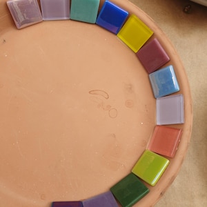 May include: A circular terracotta tray featuring a collection of square glass mosaic tiles arranged in a semi-circle along the inner rim. The tiles display a variety of colors including blue, yellow, purple, green, pink, and light lavender.