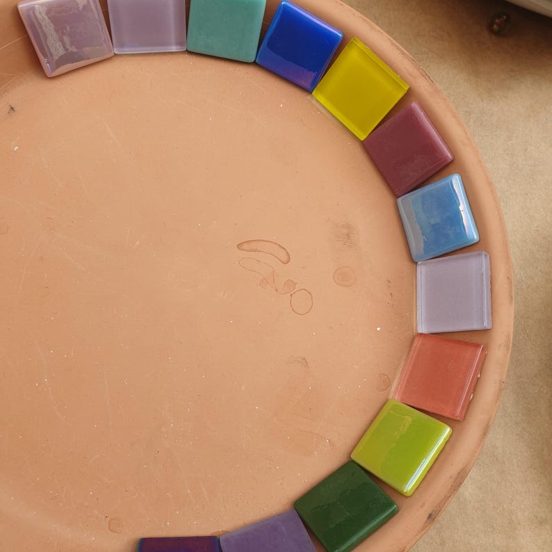 May include: A circular terracotta tray featuring a collection of square glass mosaic tiles arranged in a semi-circle along the inner rim. The tiles display a variety of colors including blue, yellow, purple, green, pink, and light lavender.