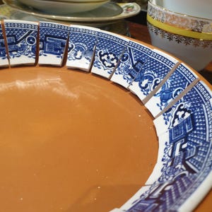 May include: A terracotta bowl featuring a decorative rim made of individual, curved ceramic shards. Each shard displays a classic blue and white willow pattern with intricate architectural and floral motifs arranged in a repeating sequence.