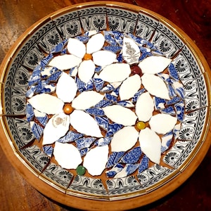 May include: A mosaic bowl with a floral design made from broken white and blue porcelain. The bowl has a wooden rim and a black and white patterned border. The mosaic includes small, colourful glass beads. The bowl's diameter is approximately 30 cm.