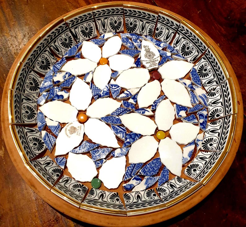 May include: A round decorative bowl featuring a mosaic design made from broken ceramic pieces. The interior displays white floral shapes with small colored centers, surrounded by blue and white patterned shards. The rim has a black and white floral border.