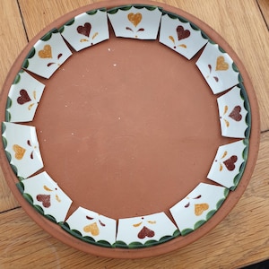 May include: A circular terracotta tray featuring a decorative border of twelve white ceramic tile segments. Each segment displays a single heart in either dark red or golden yellow, accented with small green scalloped edges.