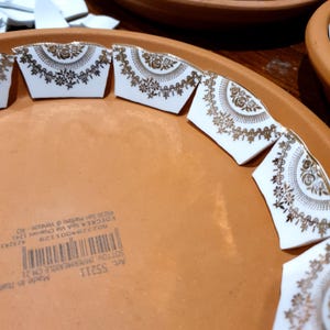May include: A terracotta saucer with a printed barcode and text including Art. 55211 and Made in Italy. The rim is decorated with white ceramic shards featuring a gold floral pattern and a scalloped edge design arranged in a circular pattern.